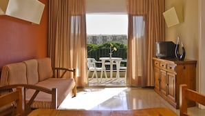 A room with a sofa, a TV on a wooden cabinet, a dining table, and a balcony with outdoor furniture. - APCOSTAS Tropik / Two Bedrooms (L'Estartit)