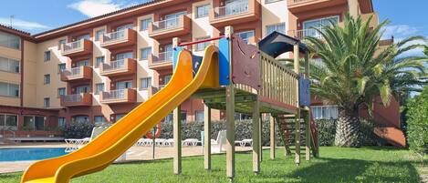 A playground with a yellow slide in front of a multi-story building with balconies.