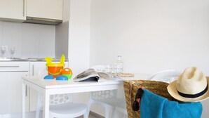 A clean, white kitchen with a dining table, chairs, and a wicker basket with a hat and sunglasses on a chair. - APCOSTAS Estartit Confort / Two Bedrooms (L'Estartit)