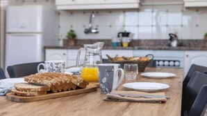 Une table de petit-déjeuner dressée avec du pain, une tasse et un pichet de jus d’orange, dans une cuisine aux armoires blanches et au dosseret carrelé.