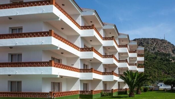 A multi-story hotel with balconies, a swimming pool, and a mountainous backdrop.