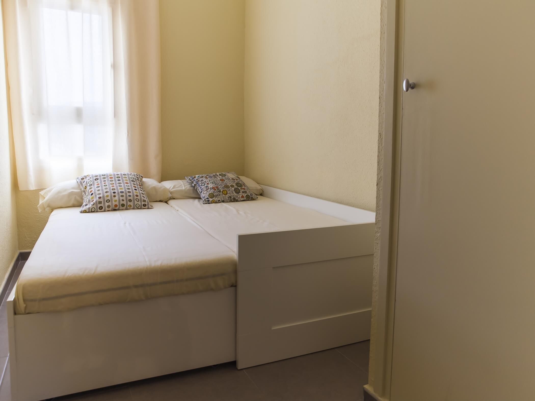 Une petite chambre avec un lit simple et un aménagement simple, comprenant un lit avec deux oreillers, une petite table de chevet et une porte.