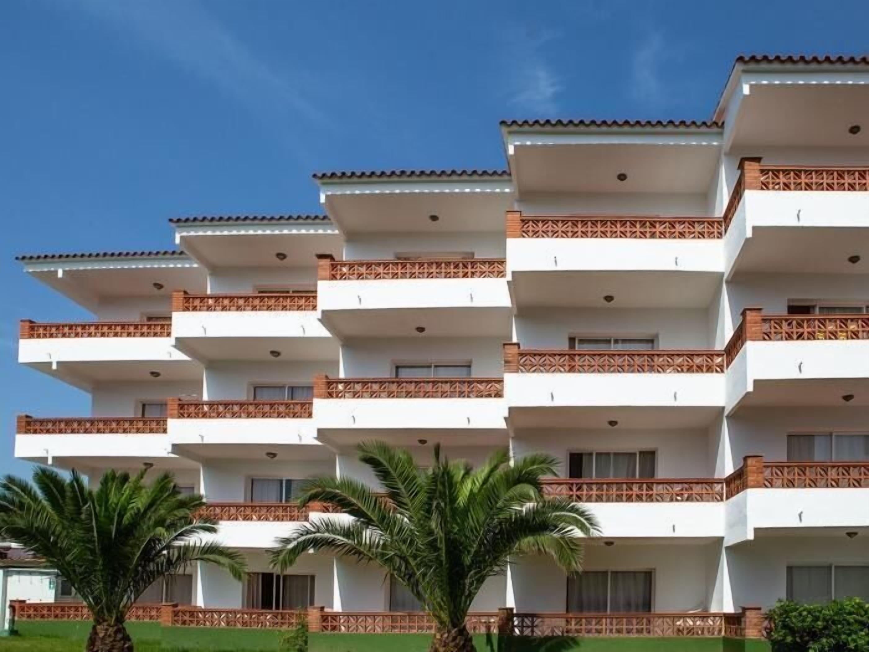 Un hôtel à plusieurs étages, doté de balcons, de palmiers et surplombant un ciel d’un bleu limpide.