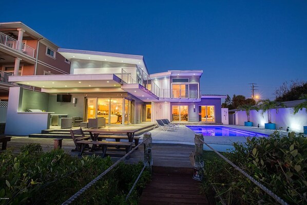 A modern two-story house with a swimming pool, outdoor seating, and a well-lit patio area.
