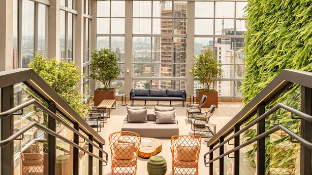 A modern interior with a sofa set, orange chairs, and a staircase leading to a balcony with a city view. - Charlie Wide Pinheiros (São Paulo)