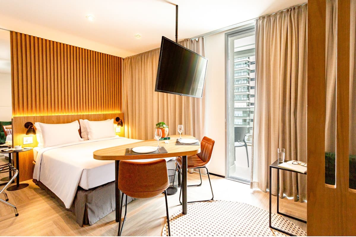 Ein Hotelzimmer mit Bett, einem Esstisch mit Stühlen, einem Fernseher und einem Balkon mit Vorhängen.