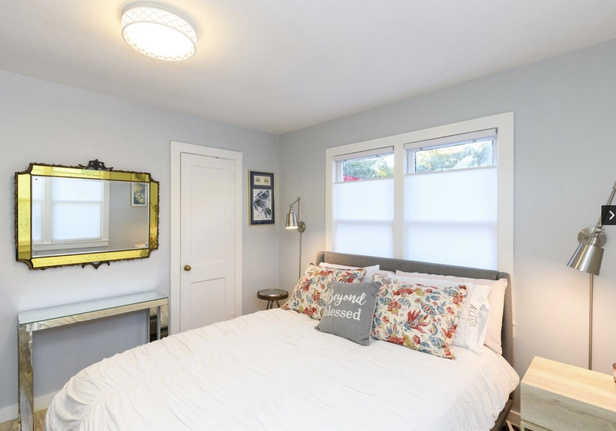 Un dormitorio con cama, espejo, puerta, ventana y mesita de noche.