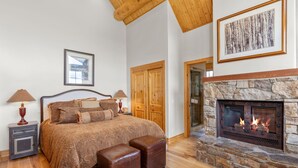 A bedroom with a large bed, a stone fireplace, wooden ceiling beams, and a framed picture on the wall.