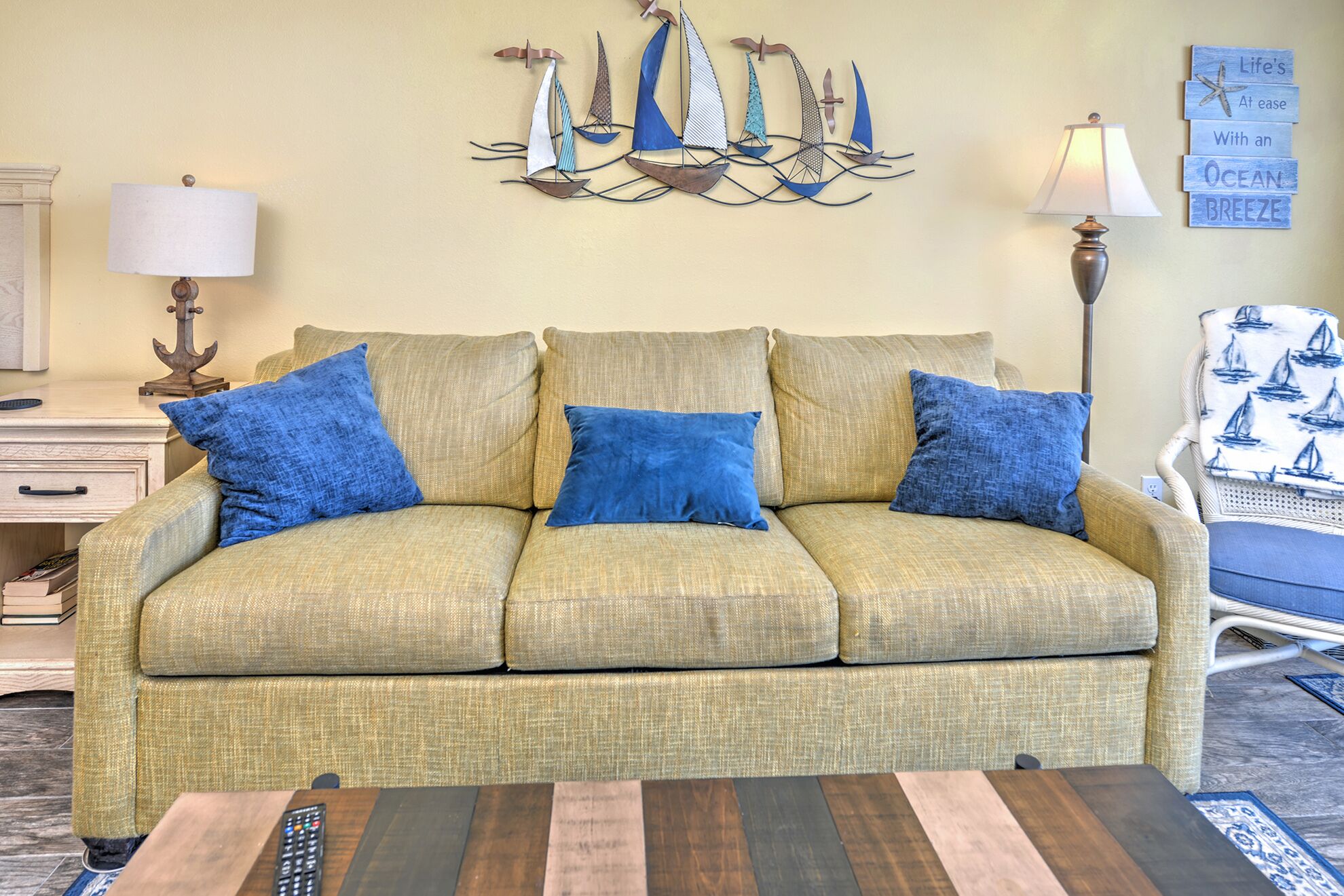 A living room with a sofa, blue cushions, a coffee table, a lamp, a wall art of sailboats, and a chair with a sailboat print.