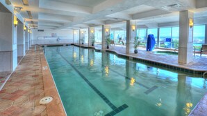 En indendørs swimmingpool med klart vand, flisegulv og udsigt til havet gennem store vinduer.