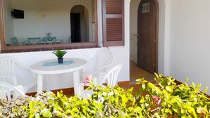 Eine kleine Terrasse mit einem runden Tisch und zwei weißen Stühlen, einem Blumentopf und Blick ins Innere durch ein Fenster mit braunen Rollläden.