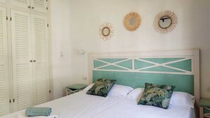 Ein ordentlich bezogenes Bett mit geometrischem Kopfteil, zwei Wanduhren und einem tropisch gestalteten Kopfkissen.