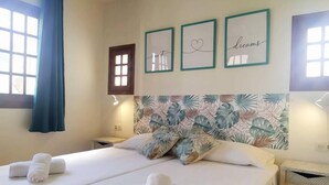 Habitación de hotel con dos camas, cabecera con estampado de hojas tropicales, cuadro en la pared y ventana con cortinas azules.