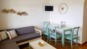 Una sala moderna con un sofá gris, sillas verde azuladas y una mesa blanca. Un televisor de pantalla plana está montado en la pared, y hay elementos decorativos en las paredes.