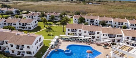 A resort with a central swimming pool, multiple white buildings with red-tiled roofs, and a view of the ocean.