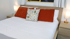 Een netjes opgemaakt bed met witte beddengoed en twee oranje kussens, een nachtkastje met een lamp, en een raam met gordijnen.