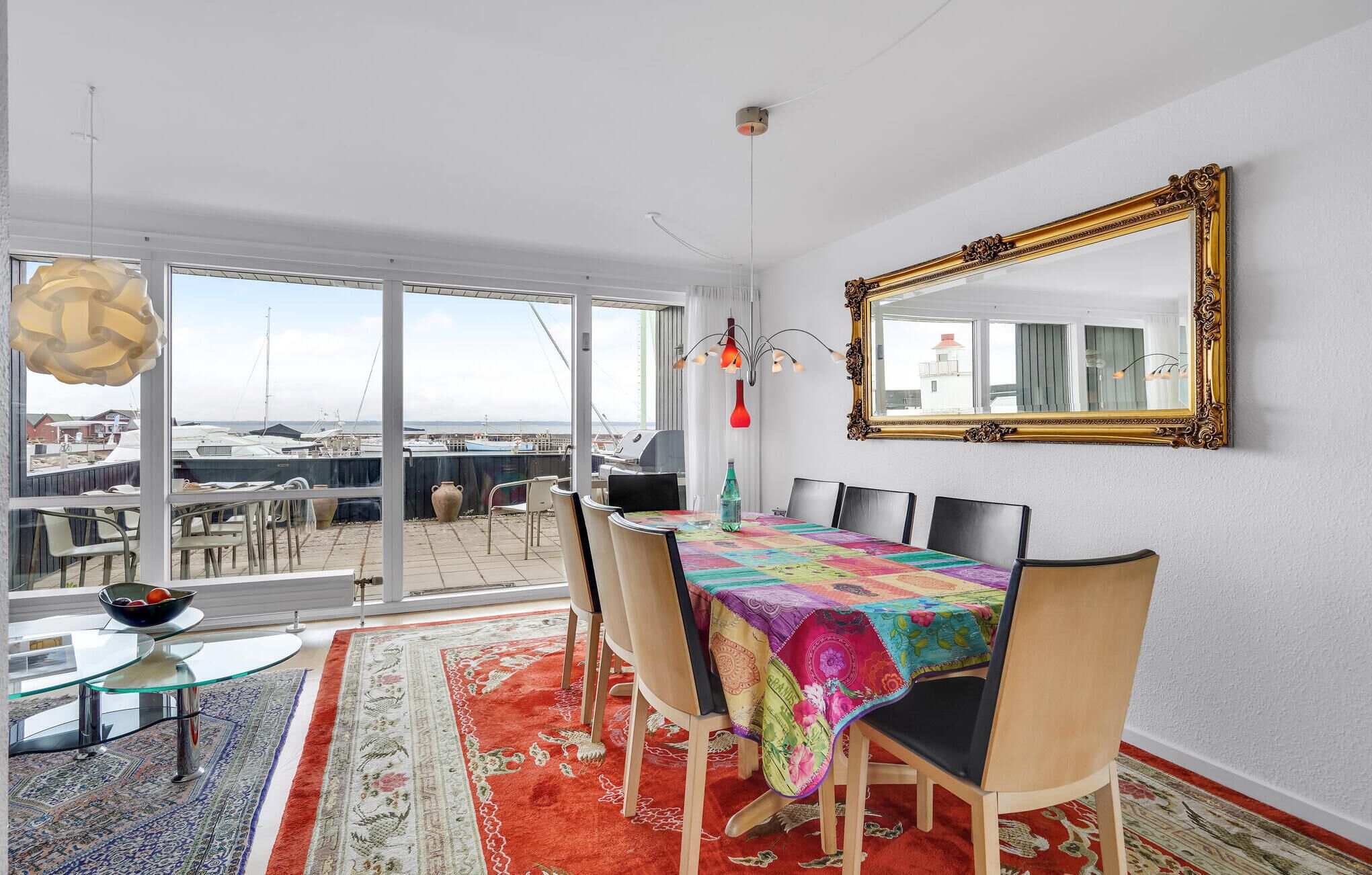 Ein Essbereich mit einem großen Tisch, Stühlen, einer bunten Tischdecke, einem Teppich, einem Glastisch und einem großen Spiegel an der Wand.