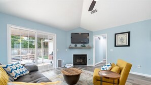 Ein Wohnzimmer mit Sofa, Sessel, Couchtisch, Kamin, Fernseher und einem großen Fenster mit Blick auf eine Terrasse.