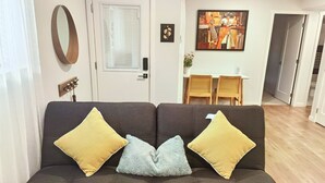 Salón con sofá marrón oscuro, tres cojines, espejo montado en la pared, puerta con ventana, cuadro, pequeña mesa con sillas y una planta.