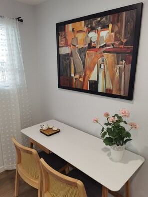 Ein Zimmer mit einem weißen Tisch, zwei Holzsesseln, einem gerahmten abstrakten Gemälde, einer Blumentopf mit rosa Blüten und einem Fenster mit durchsichtigen Vorhängen.