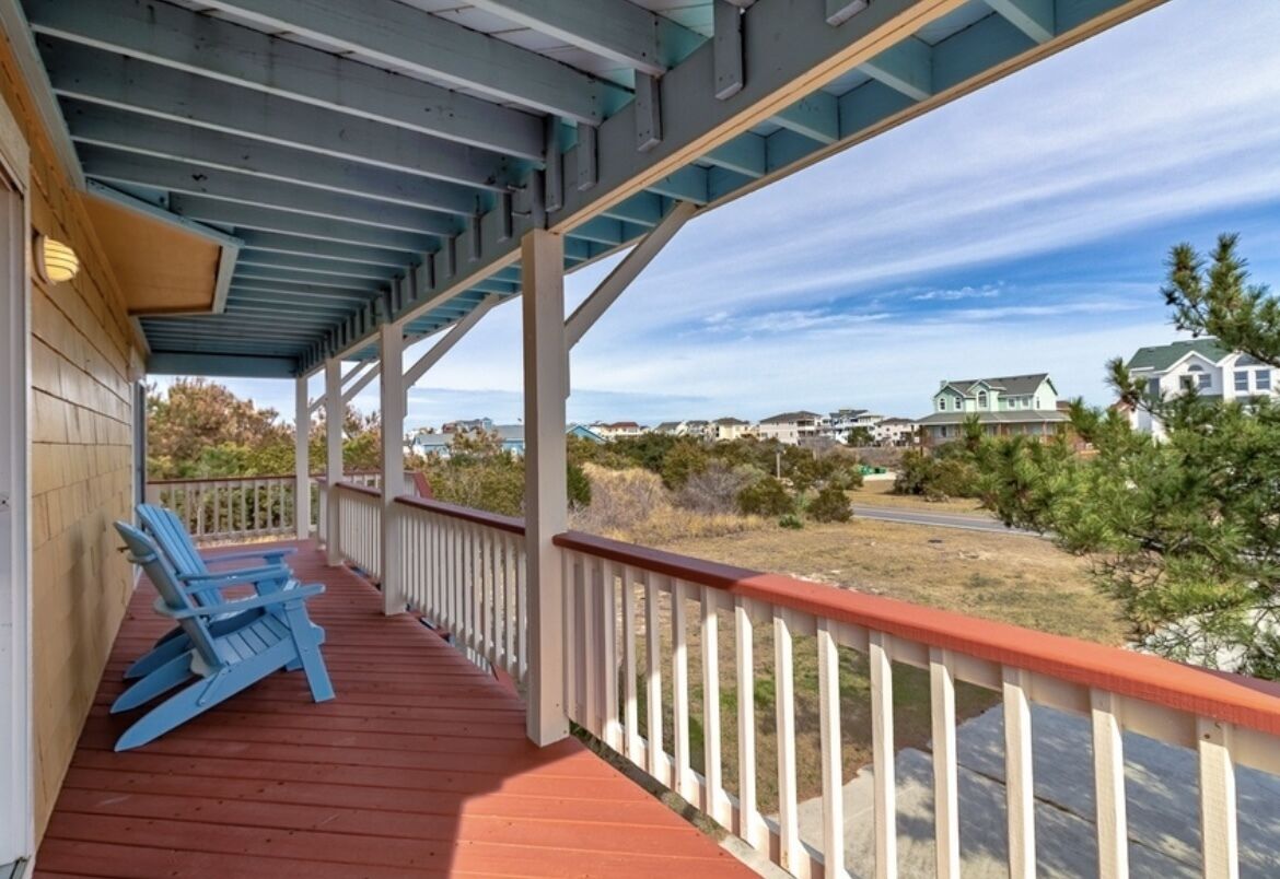 A covered porch with red flooring and white railings, overlooking a residential area with houses and a clear sky.