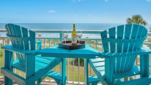 Une terrasse turquoise avec deux chaises et une table, offrant une vue sur une plage et l’océan.