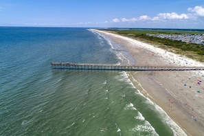 An aerial view of a beach with a pier extending into the water, a sandy shore, and a line of buildings in the distance.
