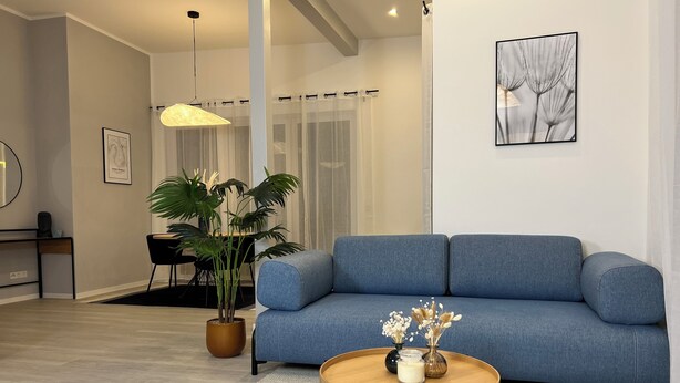 A modern living room with a blue sofa, a round coffee table, a potted plant, and a framed artwork on the wall. - Loft with wallbox, WLAN workstation, garage, for 3 people, 100 m to the Moselle (Traben-Trarbach)