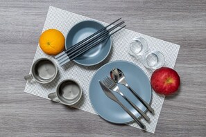 A dining setup features a blue plate, fork, spoon, chopsticks, a small bowl, and a red apple on a light wood table.