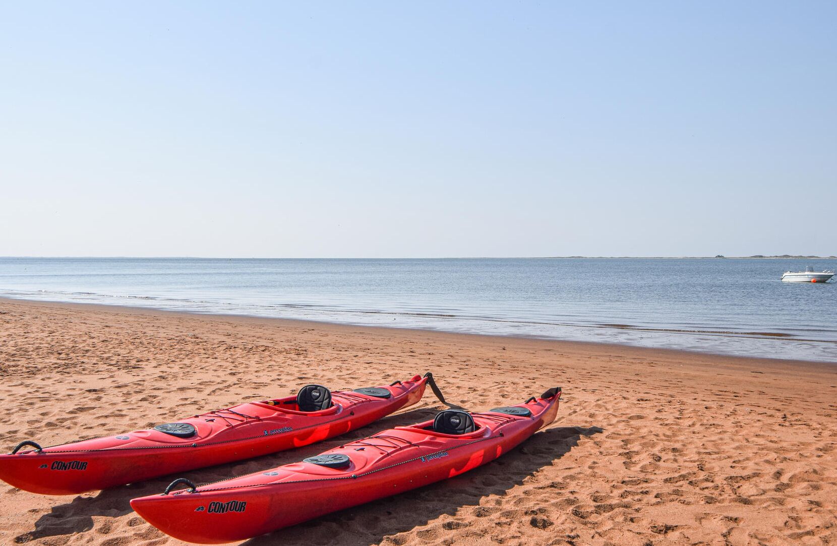 Twee rode kajaks op een zanderig strand, met rustig water en een boot in de verte.