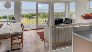 Amplo quarto com grande porta de correr de vidro que dá para uma varanda com vista para o mar. Há uma mesa de jantar de madeira com cadeiras, um sofá e uma TV sobre um suporte.