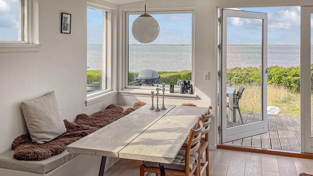 Uma sala de jantar com mesa e cadeiras, um banco com almofadas, um lustre pendente e vista para o mar através de grandes janelas.