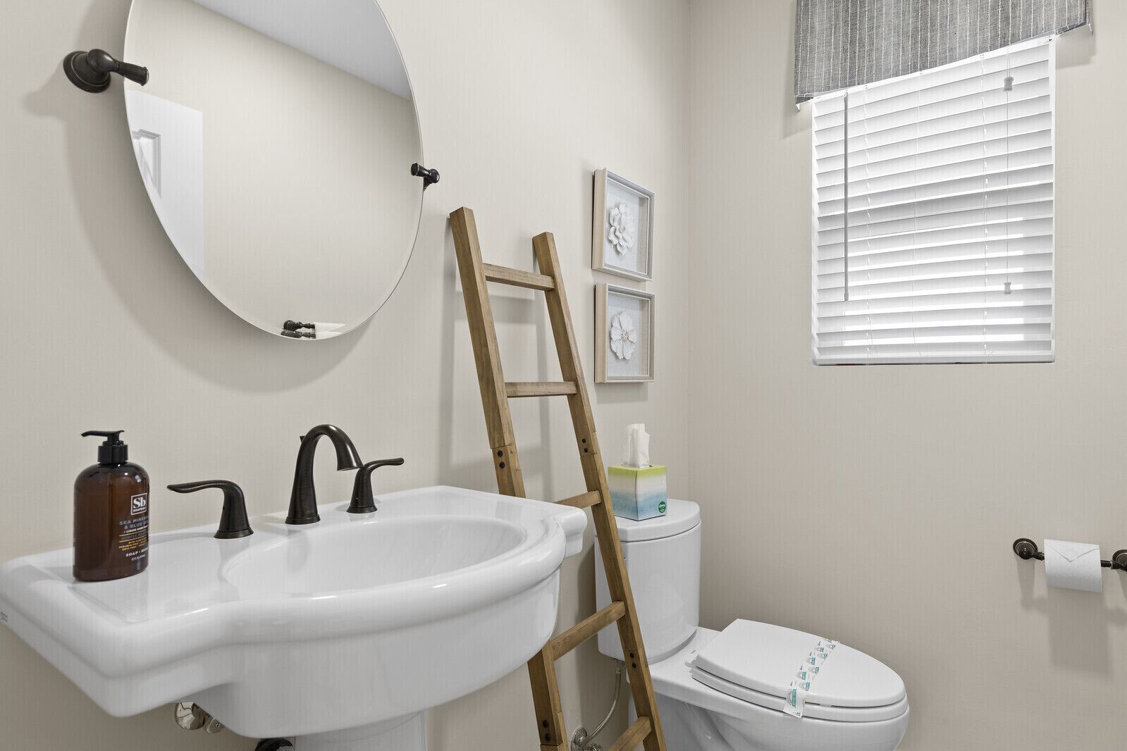 Un baño con un lavamanos blanco, dos grifos negros, una escalera de madera apoyada en la pared, un inodoro y una ventana con persianas.