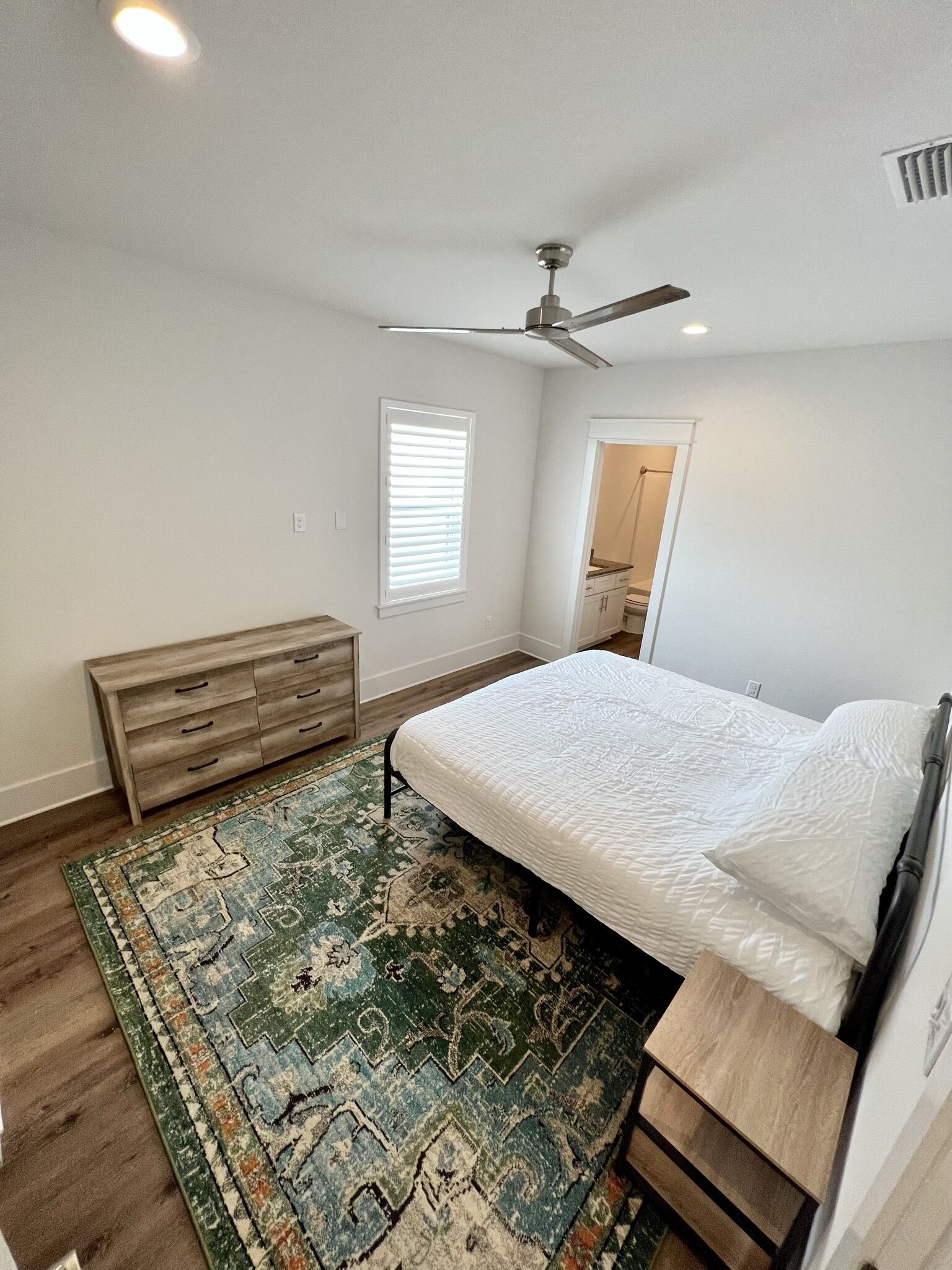 Un dormitorio con cama, ventilador de techo, ventana con persianas, banca de madera y puerta que da a un baño.