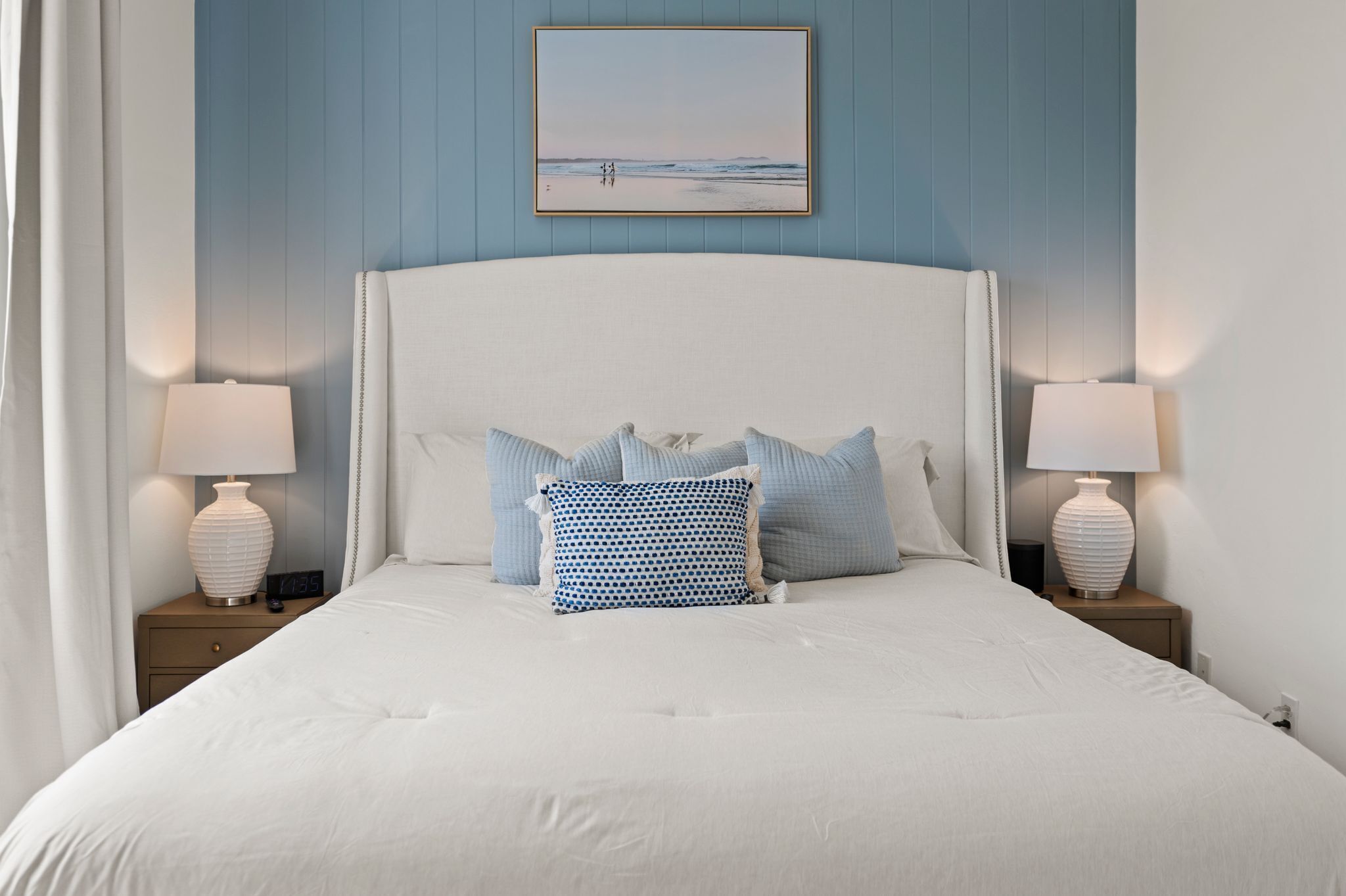 A neatly made bed with white bedding and multiple pillows, two bedside lamps, a clock, and a framed picture on the wall.