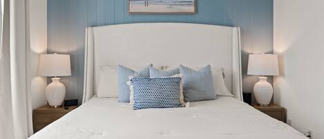 A neatly made bed with white bedding and multiple pillows, two bedside lamps, a clock, and a framed picture on the wall.