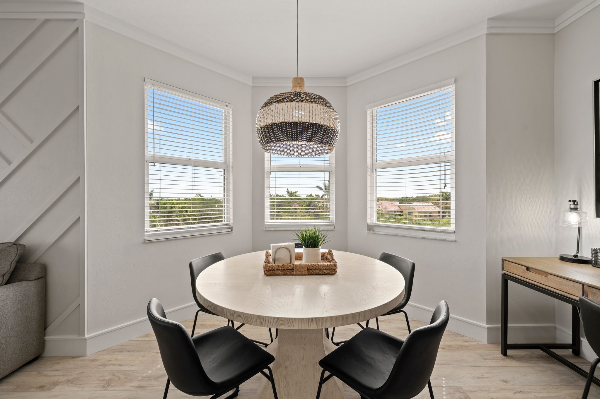 A dining area with a round table, black chairs, a hanging light fixture, and three windows with blinds.