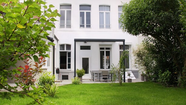 A modern building with a white facade, a front door, and a small patio area with a table and chairs. - Ligny garden (ST OMER)