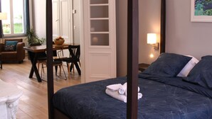 A four-poster bed with a dark blue bedspread, a white door with a glass panel, a dining area with a table and chairs, and a lamp on the wall. - Ligny garden (ST OMER)