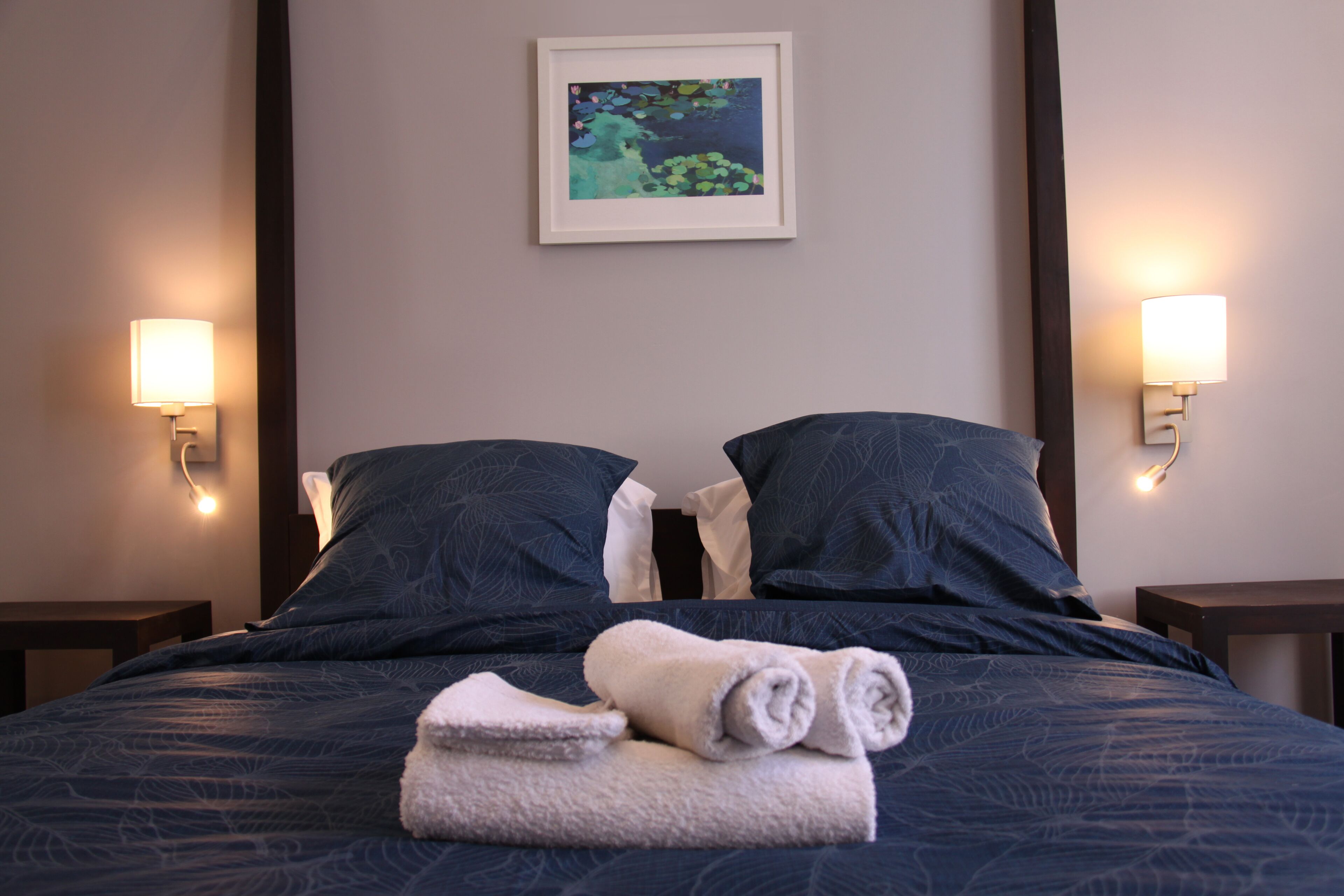 Een net opgemaakt bed met donkerblauwe lakens, twee kussens en een opgerolde witte handdoek. Aan beide kanten van het bed zijn twee wandlampen bevestigd.