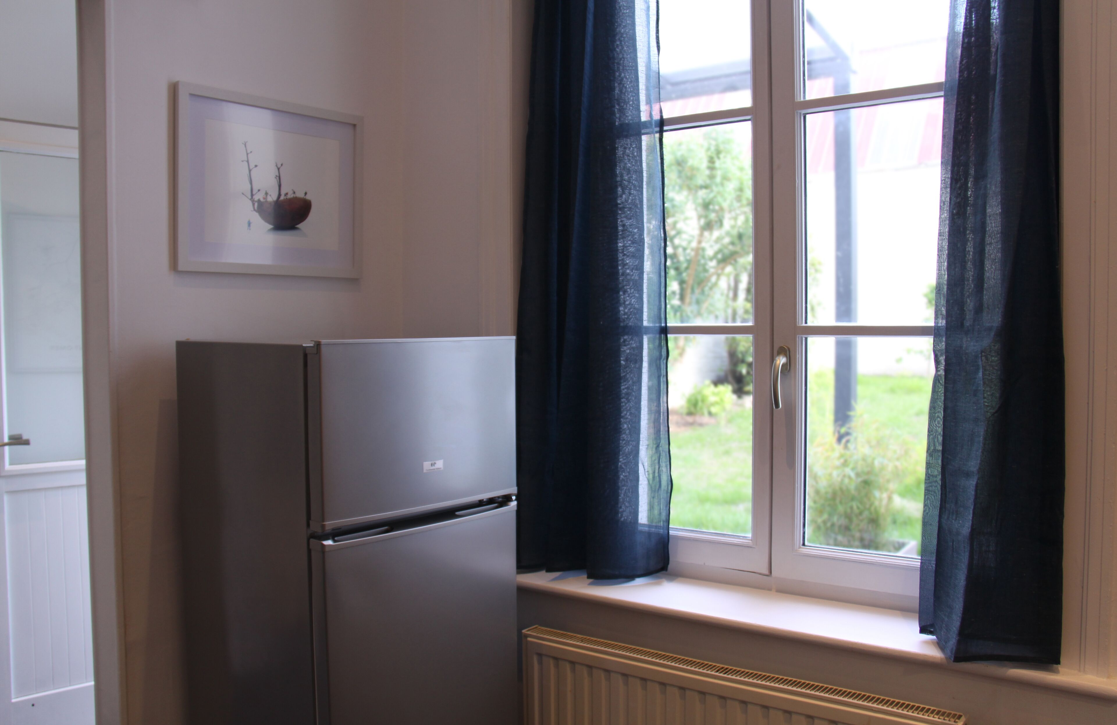 Una cocina con refrigerador, ventana con cortinas e imagen enmarcada en la pared.