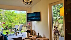 A living room with a TV, a sofa, a coffee table, and a view of a garden with chairs and a table outside.