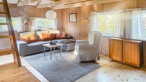 A cozy cabin interior with a wooden floor, a sofa with orange cushions, a chair, a small table, and a window with curtains. - 2 bedroom awesome home in Sejerø (Sejerø)
