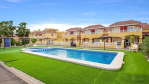 Una piscina con un motivo a mosaico geometrico, circondata da un prato curato e una fila di edifici gialli con tetti rossi e balconi.