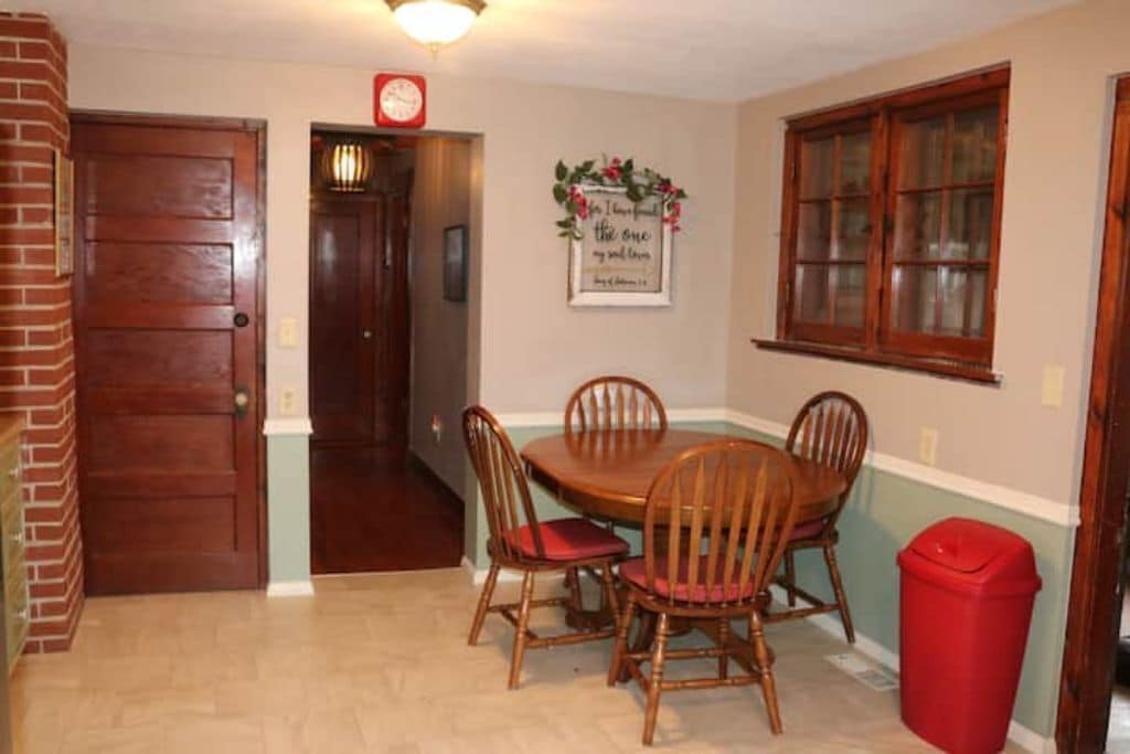 A dining area with a wooden table and four chairs, a red trash bin, and a wall clock.