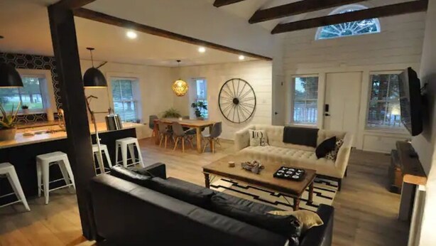 A spacious living area with a sofa, coffee table, and a dining table with chairs. There is a large wheel in the background, and the room has wooden beams on the ceiling. - Gorgeous Gem in Niagara’s Wine Country (Niagara Falls)
