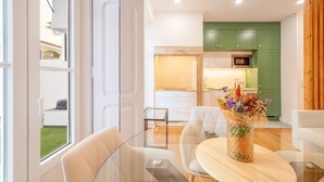 Uma sala de estar moderna com uma mesa de jantar de vidro, sofás brancos e uma cozinha verde ao fundo.