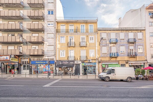 Vista da rua com edifícios altos, uma van branca estacionada na lateral e diversas lojas, incluindo um bar e uma ótica.
