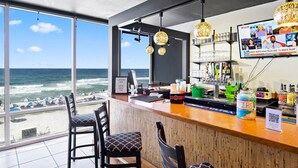 Un bar con vistas al océano, con taburetes, una barra con caja registradora y una pantalla de televisión mostrando noticias.