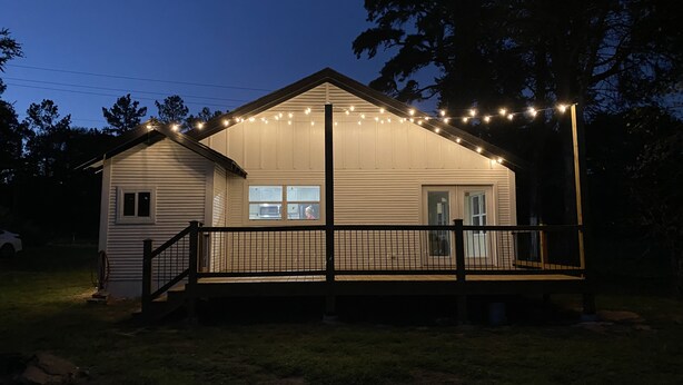 A small, single-story house with a porch and a covered area, surrounded by a fence.